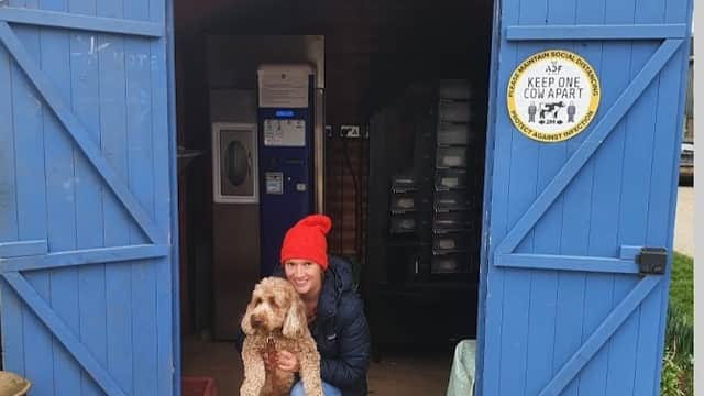 Blue shed entrance with a woman in a red hat holding a dog, surrounded by milk bottles and greenery.