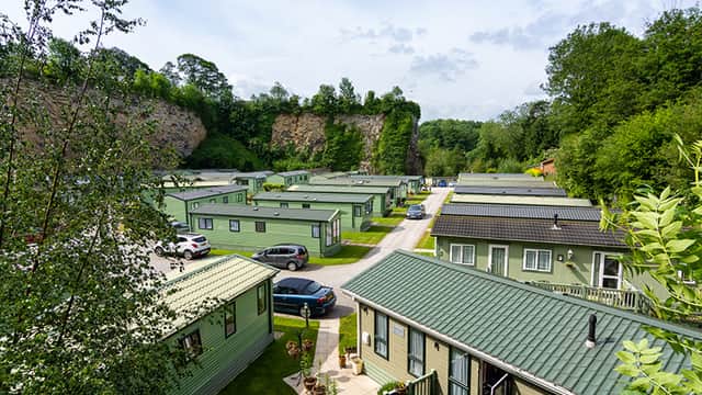 Aerial view of Low Bridge Holiday Park featuring green holiday homes surrounded by trees and a rocky cliff backdrop.