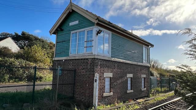 Historic railway signal box with green wooden exterior, surrounded by greenery and tracks under a blue sky.