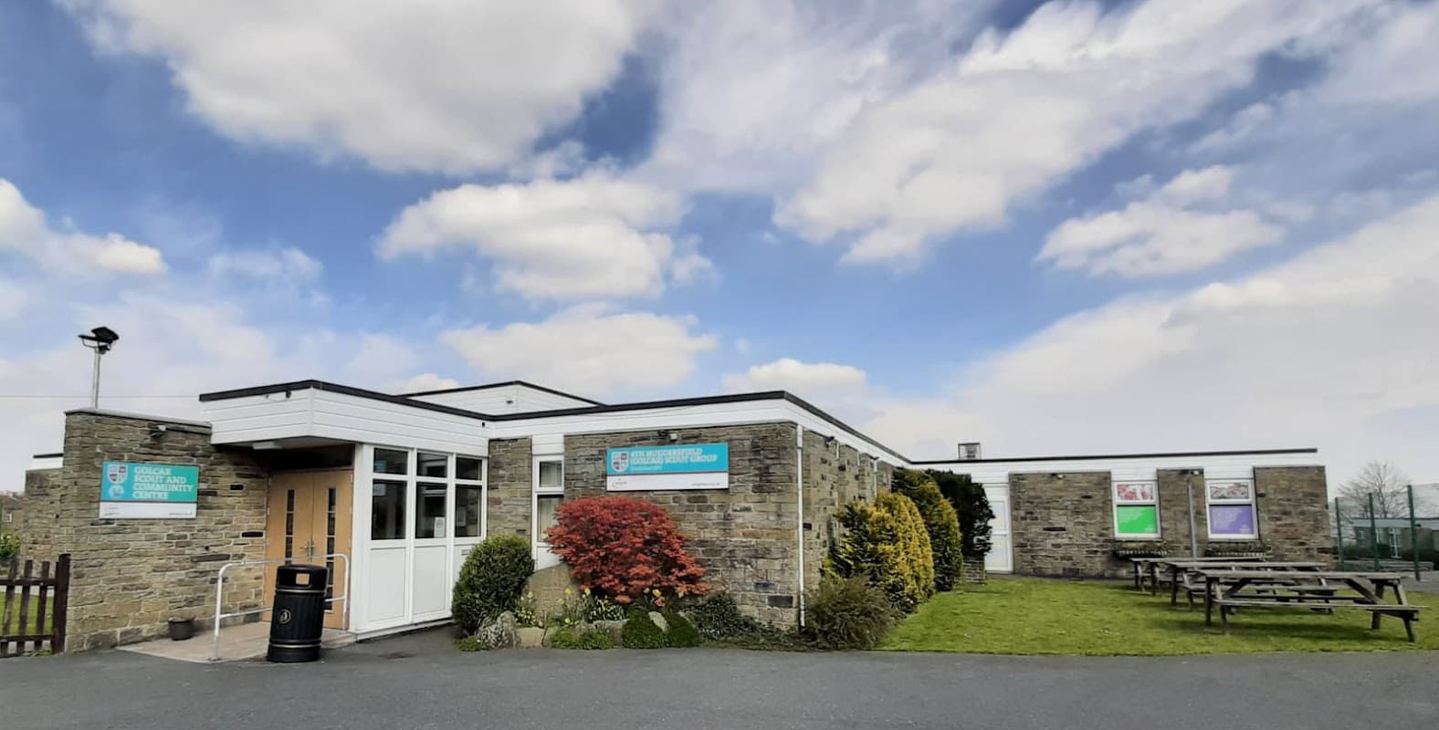 Modern stone building with large windows, green lawn, picnic tables, and a blue sky with scattered clouds.