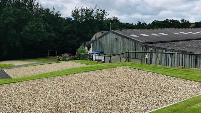 Gravel camping pitches with green grass, surrounded by trees and a rustic building under a cloudy sky.