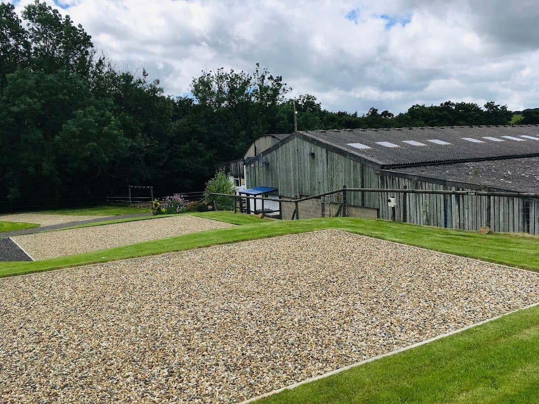 Gravel camping pitches with green grass, surrounded by trees and a rustic building under a cloudy sky.
