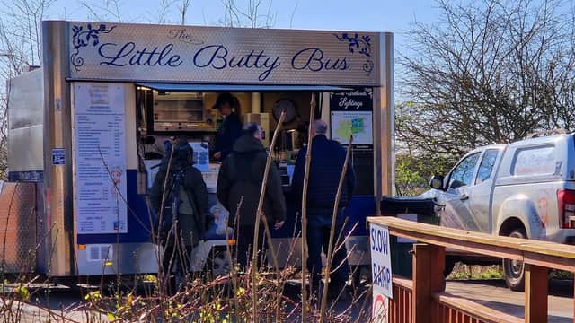 A food truck named "The Little Butty Bus" with customers ordering, surrounded by trees and a parked vehicle.