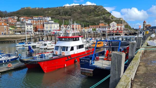 Colorful boats docked at Scarborough's West Pier, with coastal buildings and a green hillside in the background.