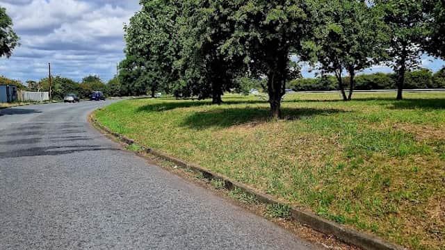 Curved road lined with trees and grassy areas, with a cloudy blue sky above in Thimbleby, North Yorkshire.