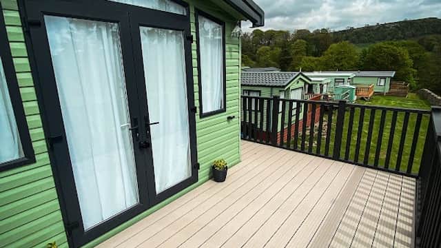 Green cabin with large glass doors on a deck, overlooking grassy areas and other cabins under a cloudy sky.