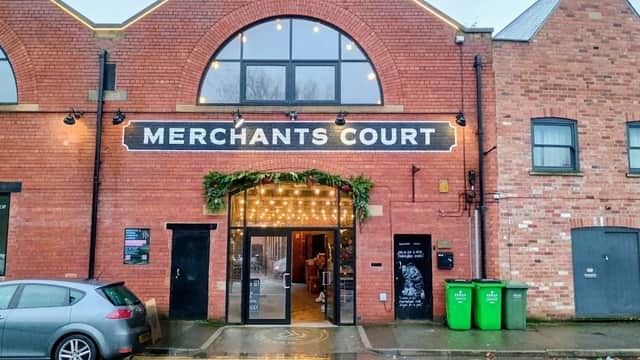 Brick building entrance with "MERCHANTS COURT" sign, festive lights, and green bins nearby. Wet pavement reflects light.