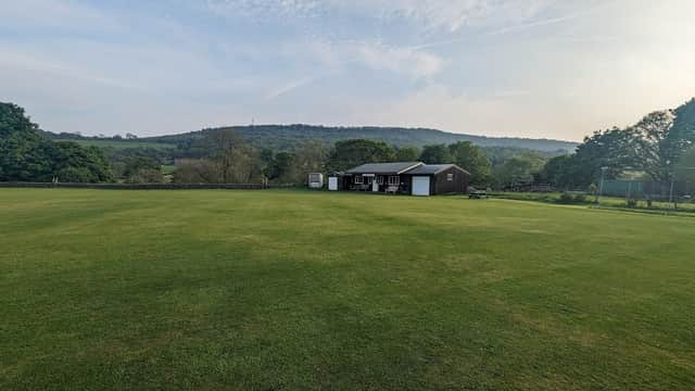 Green cricket field with a clubhouse and trees, set against a backdrop of hills and a clear blue sky.