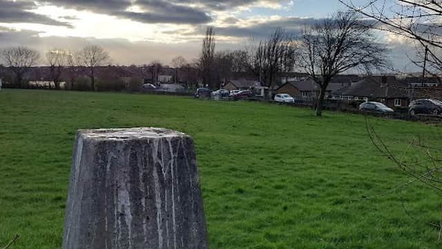 Concrete marker in grassy field under a cloudy sky, with trees and houses visible in the background.