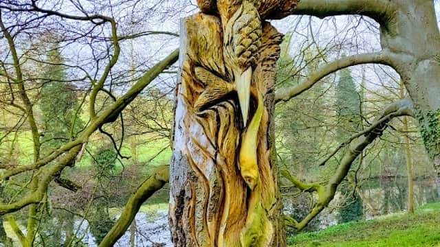 Wooden sculpture of a bird perched on a tree stump, surrounded by lush greenery and a pond in the background.
