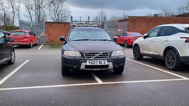 A black car parked across two spaces in a Pay & Display car park, with other cars and a cloudy sky in the background.