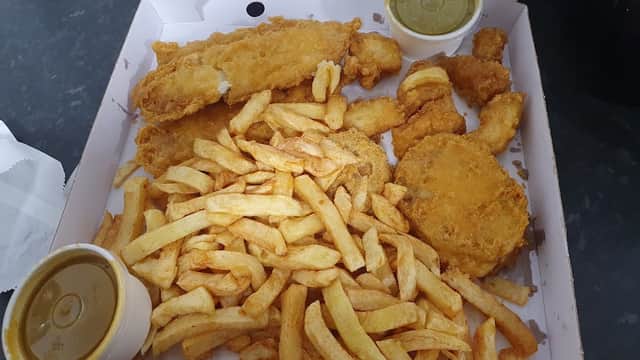 A takeaway box filled with golden fried fish, chicken pieces, and a generous portion of chips, accompanied by two sauces.