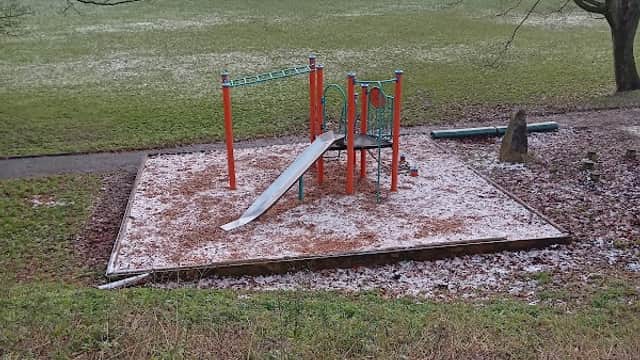 Playground with a slide and climbing frame, surrounded by grass and trees, lightly dusted with snow.