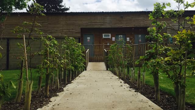 Pathway lined with young trees leading to a wooden building at The Burrows Country Park, surrounded by green grass.