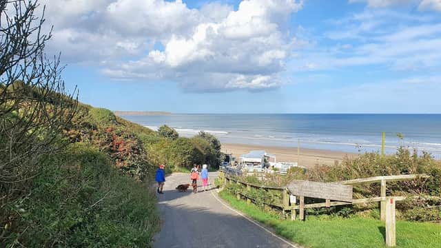 Scenic view of a coastal road leading to a beach, with people walking and lush greenery on either side.