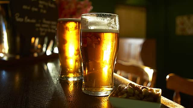 Two pints of golden beer on a wooden bar, with a bowl of snacks and a dark green pub interior in the background.