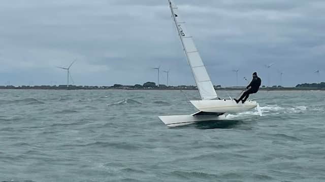 A sailor on a small dinghy navigating choppy waters, with wind turbines visible in the background under a cloudy sky.