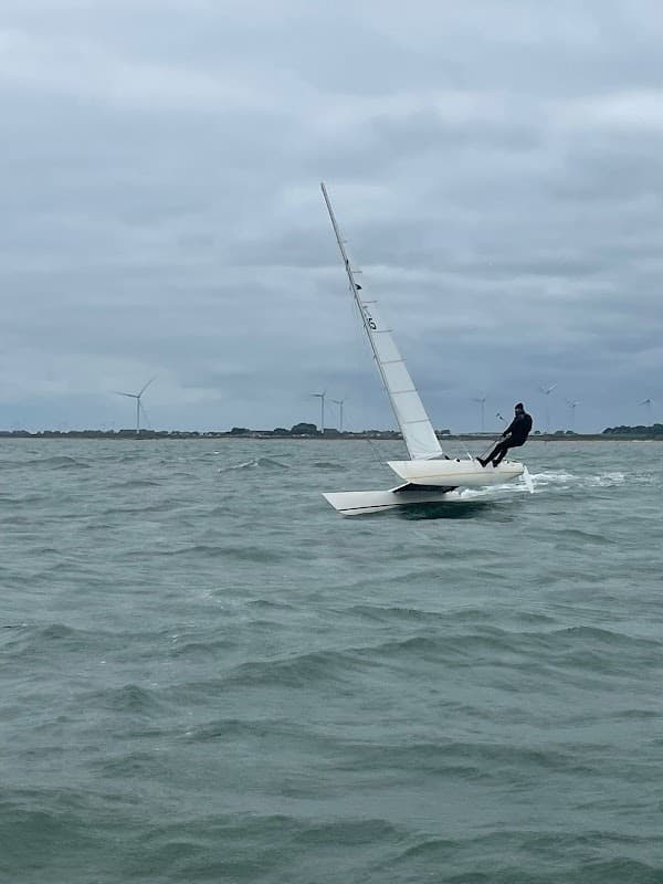 A sailor on a small dinghy navigating choppy waters, with wind turbines visible in the background under a cloudy sky.