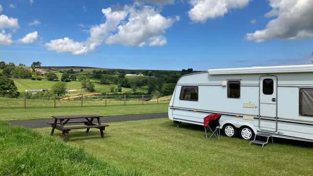 Caravan parked on green grass with picnic table, surrounded by rolling hills and a blue sky with fluffy clouds.
