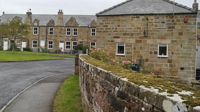 Stone building with a sloped roof, surrounded by greenery and a moss-covered wall, in a quaint village setting.