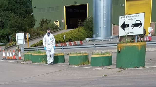 Household waste and recycling site with green barriers, signage, and a worker in protective gear walking on the pavement.