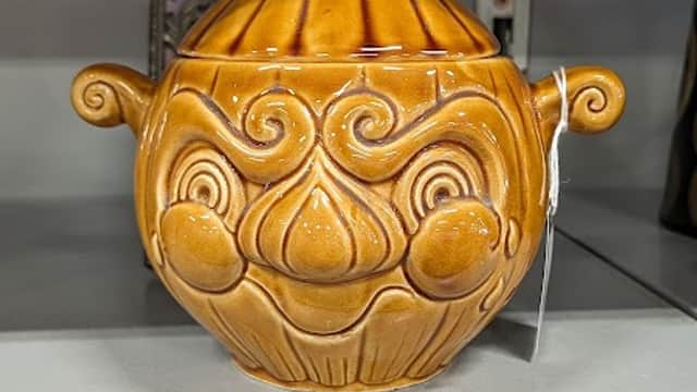 Brown ceramic pot with a whimsical face design, featuring a rounded body and a narrow neck, displayed on a shelf.
