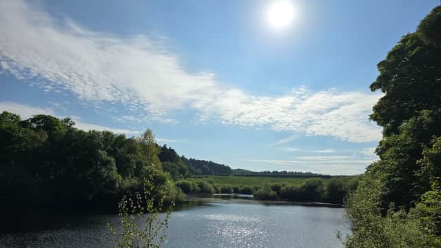 Bright sun above a serene reservoir surrounded by lush greenery and rolling hills under a partly cloudy sky.