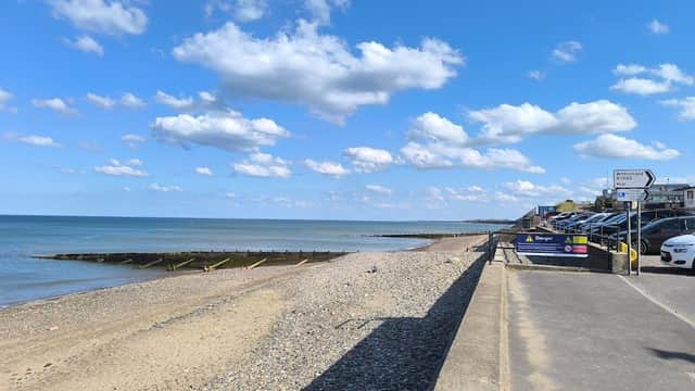 Coastal view with a sandy beach, pebbles, calm sea, and a car park lined with vehicles under a blue sky.