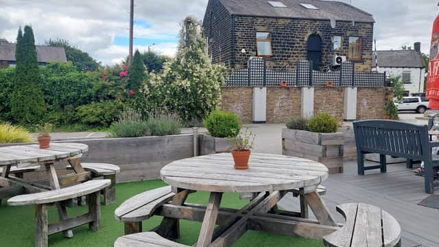 Wooden picnic tables on green grass, surrounded by flowers and shrubs, with a stone building in the background.