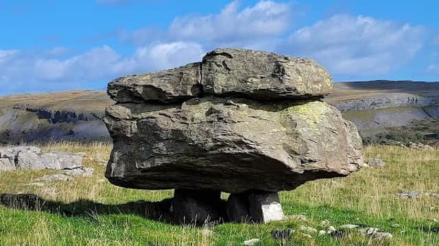 Large, flat boulder balanced on smaller stones, set against a blue sky and grassy landscape in Austwick, Yorkshire.
