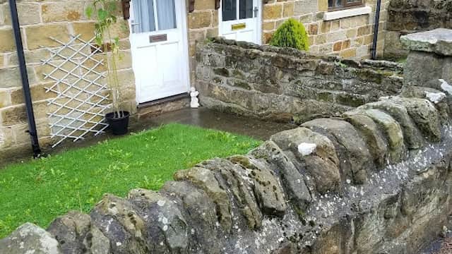 Two cottage doors with wooden canopies, surrounded by a stone wall and a small green lawn in Cloughton, Yorkshire.