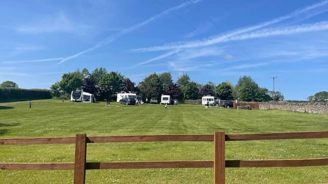 Green lawn with caravans parked, surrounded by trees under a clear blue sky with wispy clouds. Wooden fence in foreground.