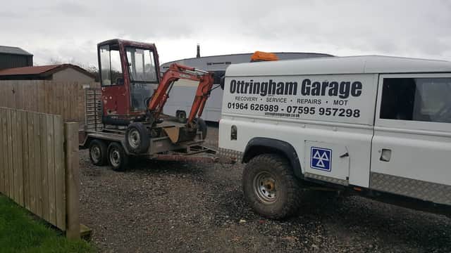 Red mini excavator on a trailer next to a service vehicle with "Ottringham Garage" branding. Gravel surface and cloudy sky.
