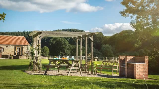 Rustic wooden pergola with picnic table and chairs in a grassy area, surrounded by trees and a brick barbecue.