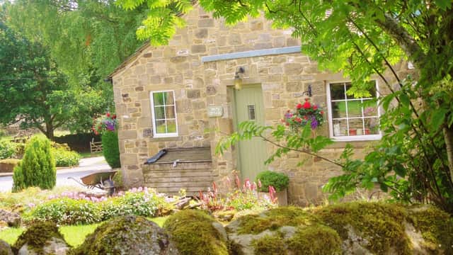 Charming stone cottage with green door, flower boxes, and lush garden, set against a backdrop of trees and a mossy wall.
