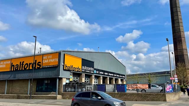 Halfords store with bright signage, cars parked outside, and a tall industrial chimney in the background under a cloudy sky.