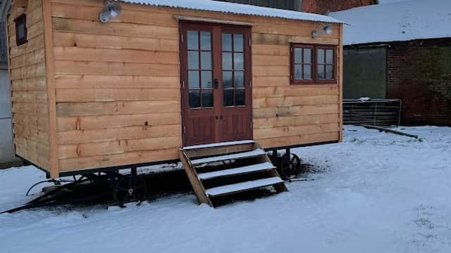 Wooden cabin on wheels in a snowy field, surrounded by farm buildings under a cloudy sky.
