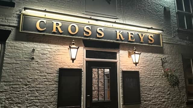 Illuminated sign reading "CROSS KEYS" on a brick pub exterior with two wall-mounted lanterns at night.