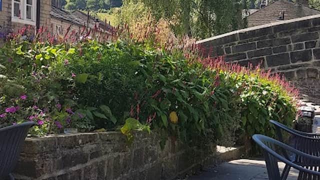 Lush greenery and colorful flowers surround a stone wall, with blue sky and trees in the background. Outdoor seating visible.