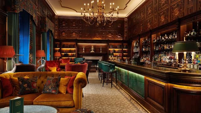 Elegant bar with ornate wood paneling, plush seating, and a well-stocked bar, illuminated by chandeliers.