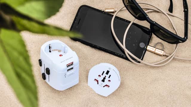 White travel adapter, smartphone, earphones, and sunglasses on sandy surface with green leaves in the corner.