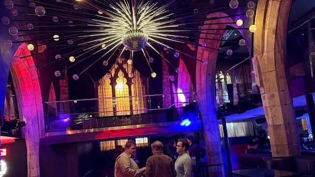 A stylish pub interior with arched stone walls, a large chandelier, and patrons conversing in a vibrant atmosphere.