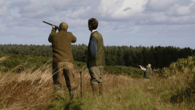 Two men in shooting attire aim shotguns in a grassy field, with trees and a cloudy sky in the background.
