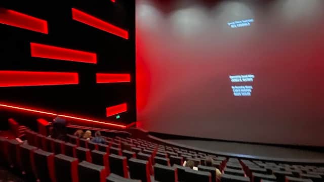 Modern cinema interior with red accents, empty rows of plush black seats, and a large blank screen at the front.