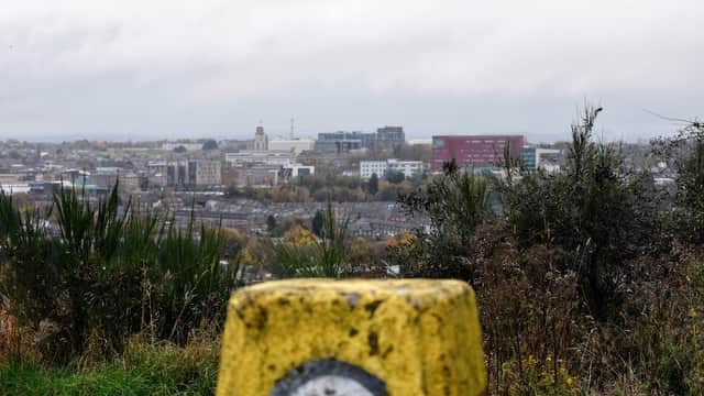 Yellow trig point in foreground with a panoramic view of a town and cloudy sky in the background.