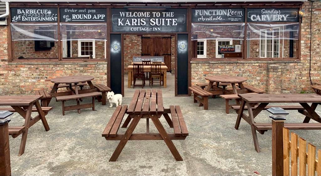 Welcoming entrance of The Cottage Inn with outdoor seating, a white dog, and signage for events and dining.
