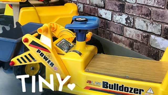 Bright yellow toy bulldozer with a wooden seat, construction helmets, and safety cones against a brick wall backdrop.