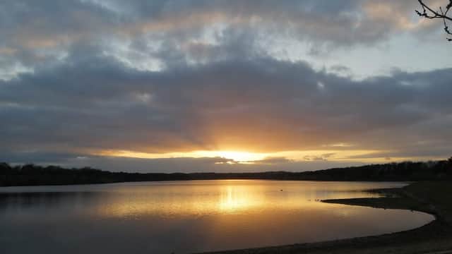 Sunset over Eccup Reservoir, with calm water reflecting clouds and a golden sky, framed by trees on the shore.