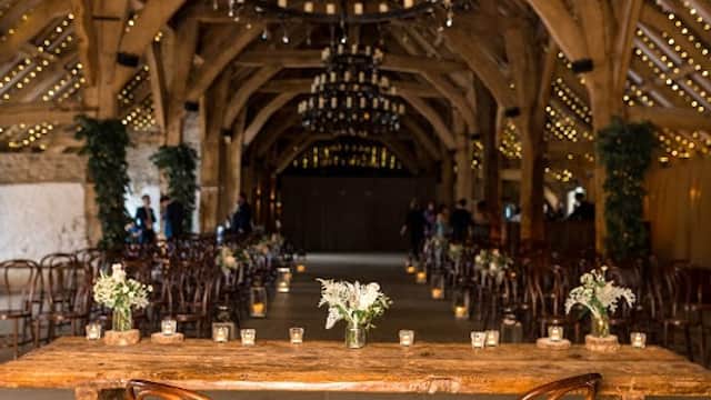 Rustic venue with wooden beams, chandeliers, and floral centerpieces on a long table, surrounded by chairs.
