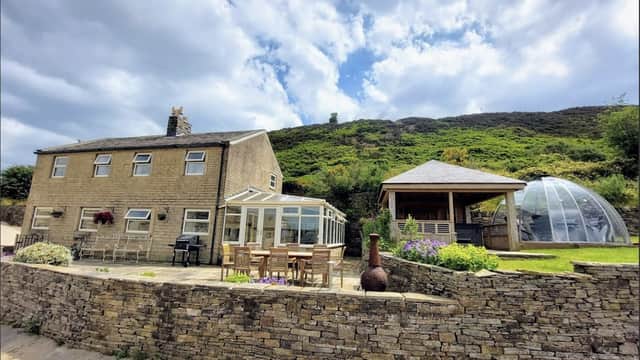 Stone farmhouse with a glass conservatory, outdoor seating, and a dome structure, set against a hilly backdrop.